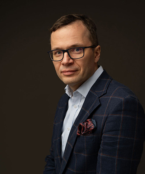 mężczyzna w okularach i garniturze pozujący do zdjęcia na jednolitym ciemnym tle