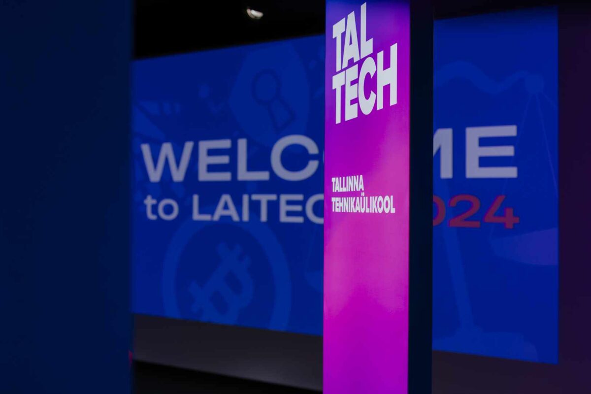 Stojący ekran z napisem TALTECH na niebiesko-fioletowym tle, w tle tło sceny konferencyjnej/