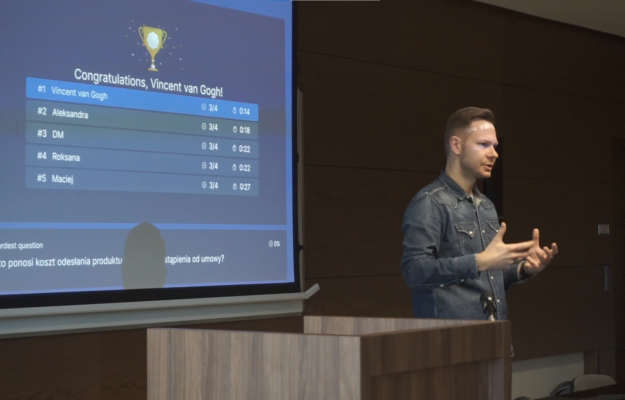 Wojciech Matelski, prezentujący na tle ekranu, na którym wyświetlany jest fragment quizu dla studentów.