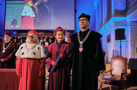 Grupa osób w strojach akademickich podczas formalnej uroczystości. Na pierwszym planie znajdują się trzy osoby. W tle widać publiczność oraz duży ekran pokazujący inne osoby w podobnych strojach akademickich.