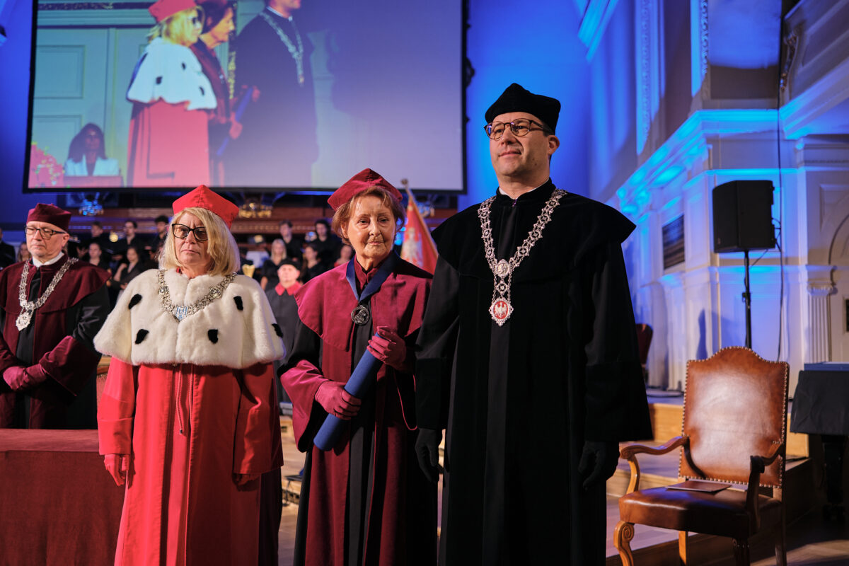 Grupa osób w strojach akademickich podczas formalnej uroczystości. Na pierwszym planie znajdują się trzy osoby. W tle widać publiczność oraz duży ekran pokazujący inne osoby w podobnych strojach akademickich.