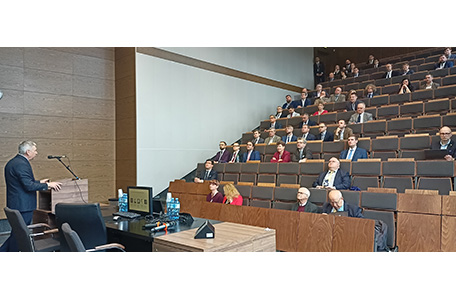 Duża grupa ludzi siedzi w sali wykładowej, zwrócona w stronę prelegenta na scenie