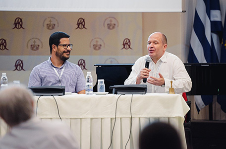 Dwaj mężczyźni siedzą przy stole, jeden z mikrofonem, prowadząc wykład