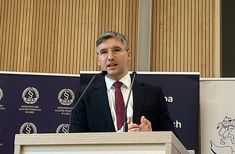 mężczyzna w garniturze i krawacie stoi przy mównicy, wygłaszając przemówienie